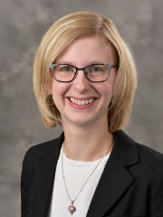 Rebecca Mushtare poses for a headshot in front of a light grey background. She wears square glasses, with a white top and black blazer. 